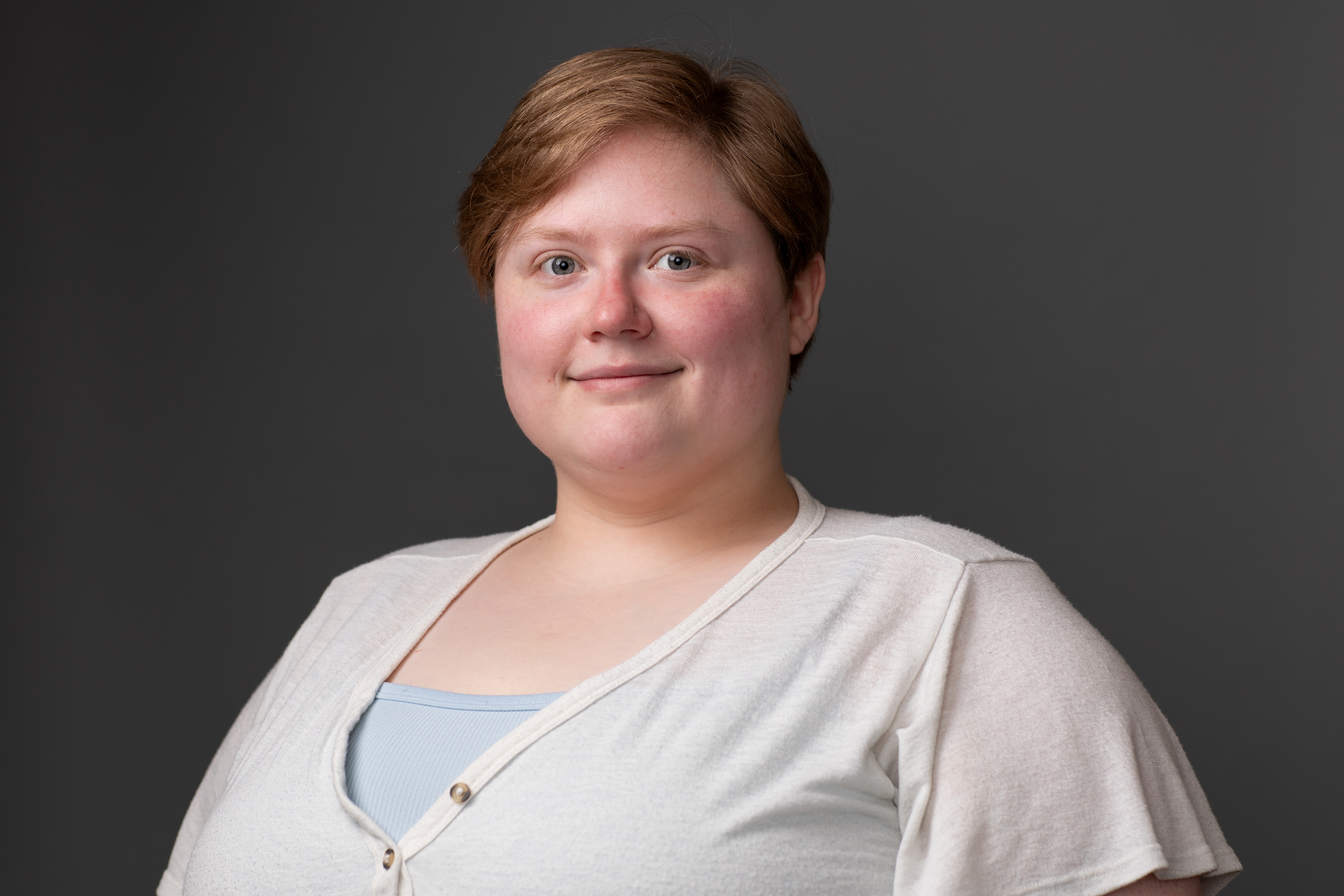 Jacquelyn's headshot. An image of a feminine person with short dirty blonde hair, wearing a white shirt that has a blue undershirt peeking through. They are looking towards the camera with a confident and warm closed mouth smile.